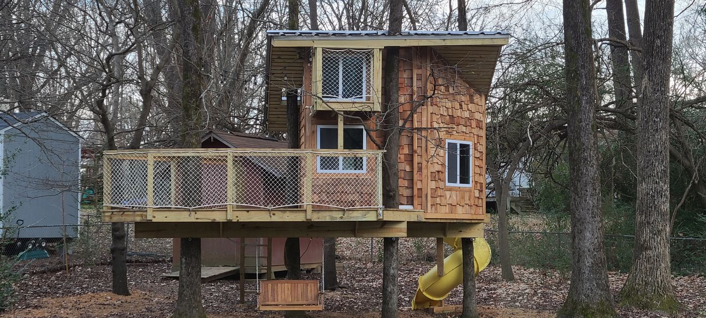 Finished two-story treehouse with cedar-shingle siding, a railed deck, a swing and a yellow slide